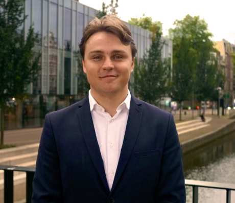 Een man in zakelijke kleding staat glimlachend buiten bij een waterkant in de stad.