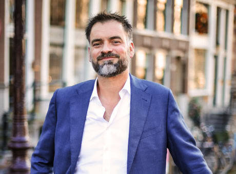 Een man in een blauwe pak en wit overhemd glimlacht in een stadsomgeving.